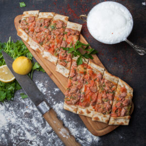 top view pide with pieces of meat and parsley and lemon and knife and ayran in cutting board on stone background
